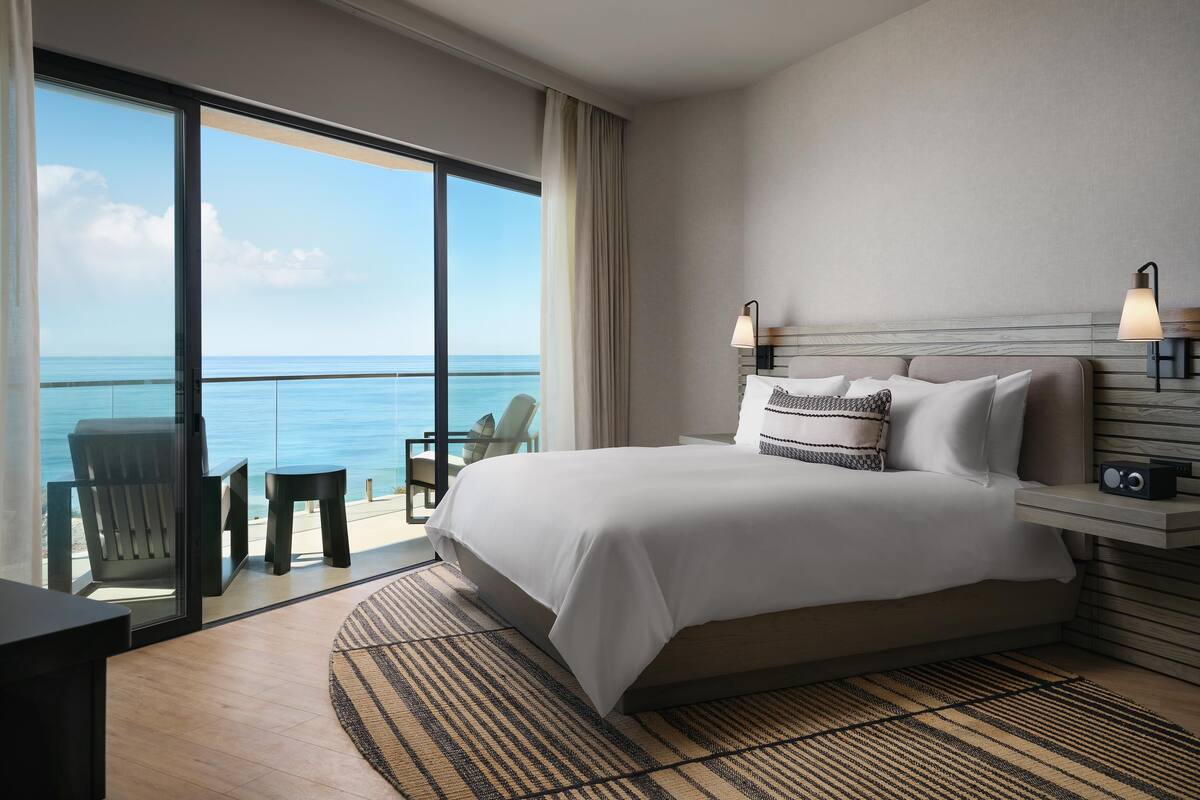 A hotel room with a large bed, two bedside lamps, a nightstand, a radio, a chair, and a view of the ocean.