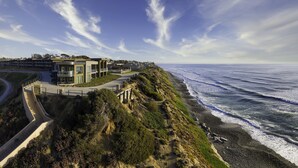 A coastal hotel with a modern design, featuring a curved driveway leading to the entrance, a beachfront view, and a rocky shoreline.