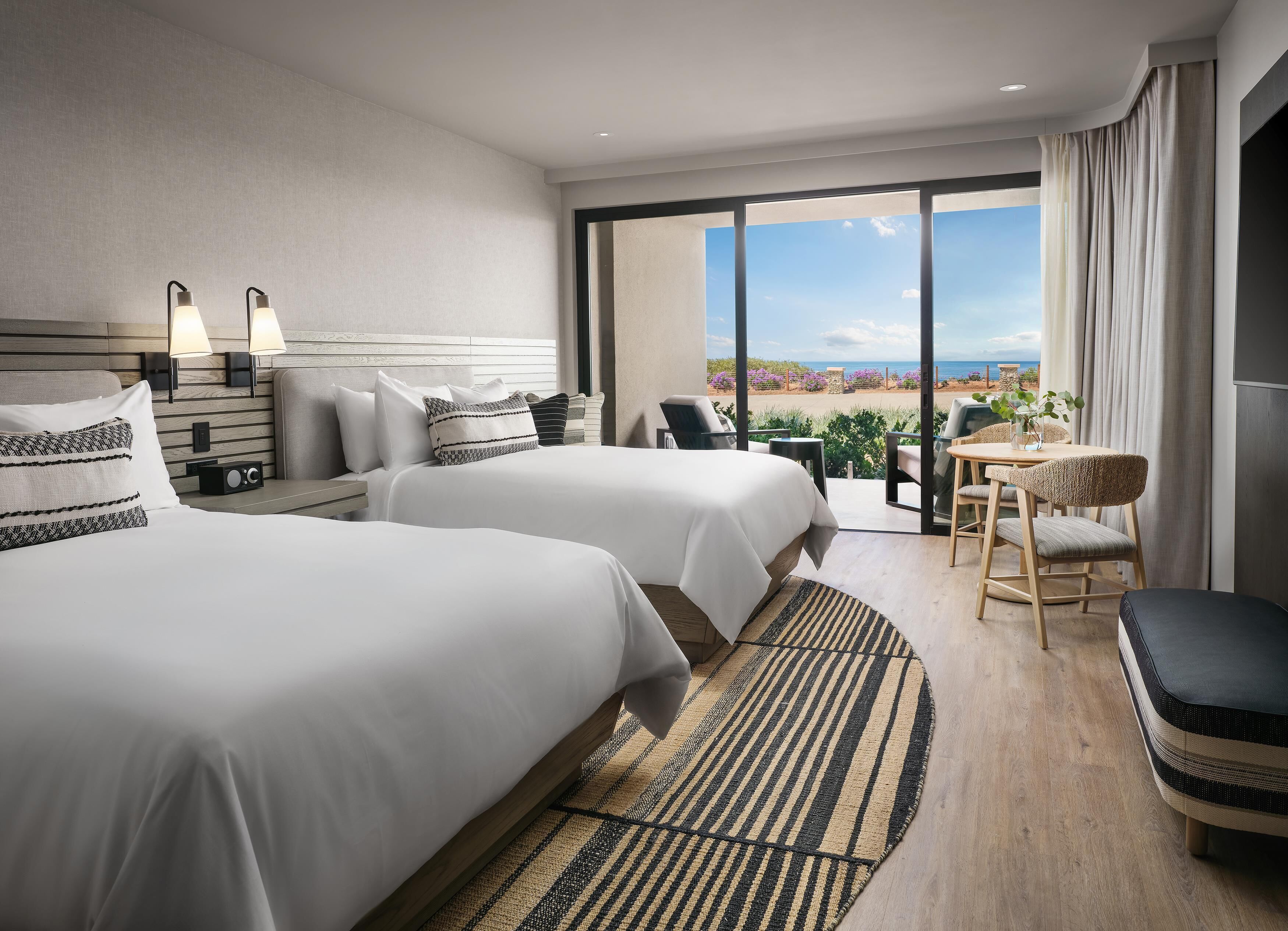 A hotel room with two beds, a balcony with a view, a small table, and a chair.