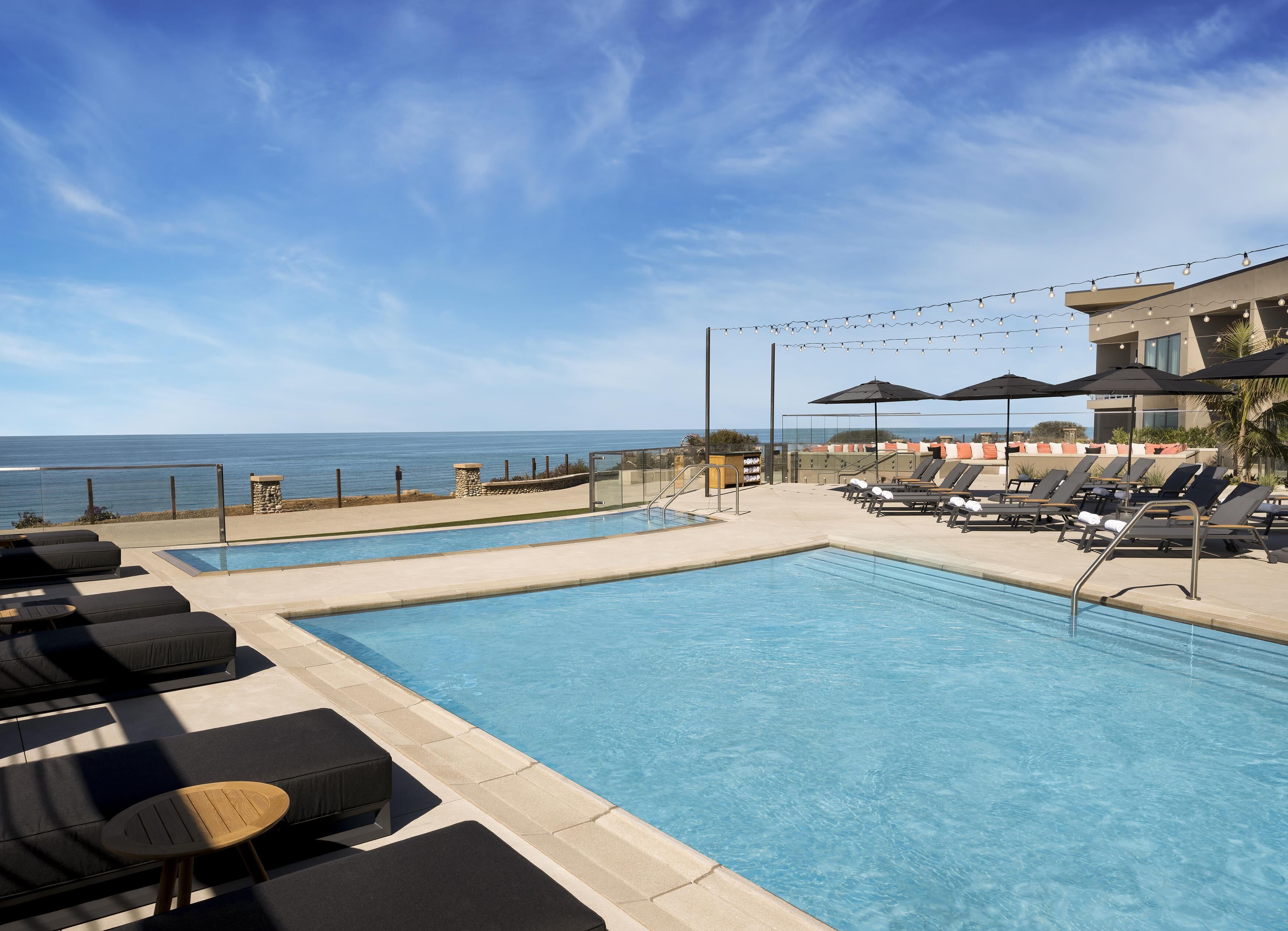 A pool area with lounge chairs, a small table, and umbrellas, overlooking the ocean.