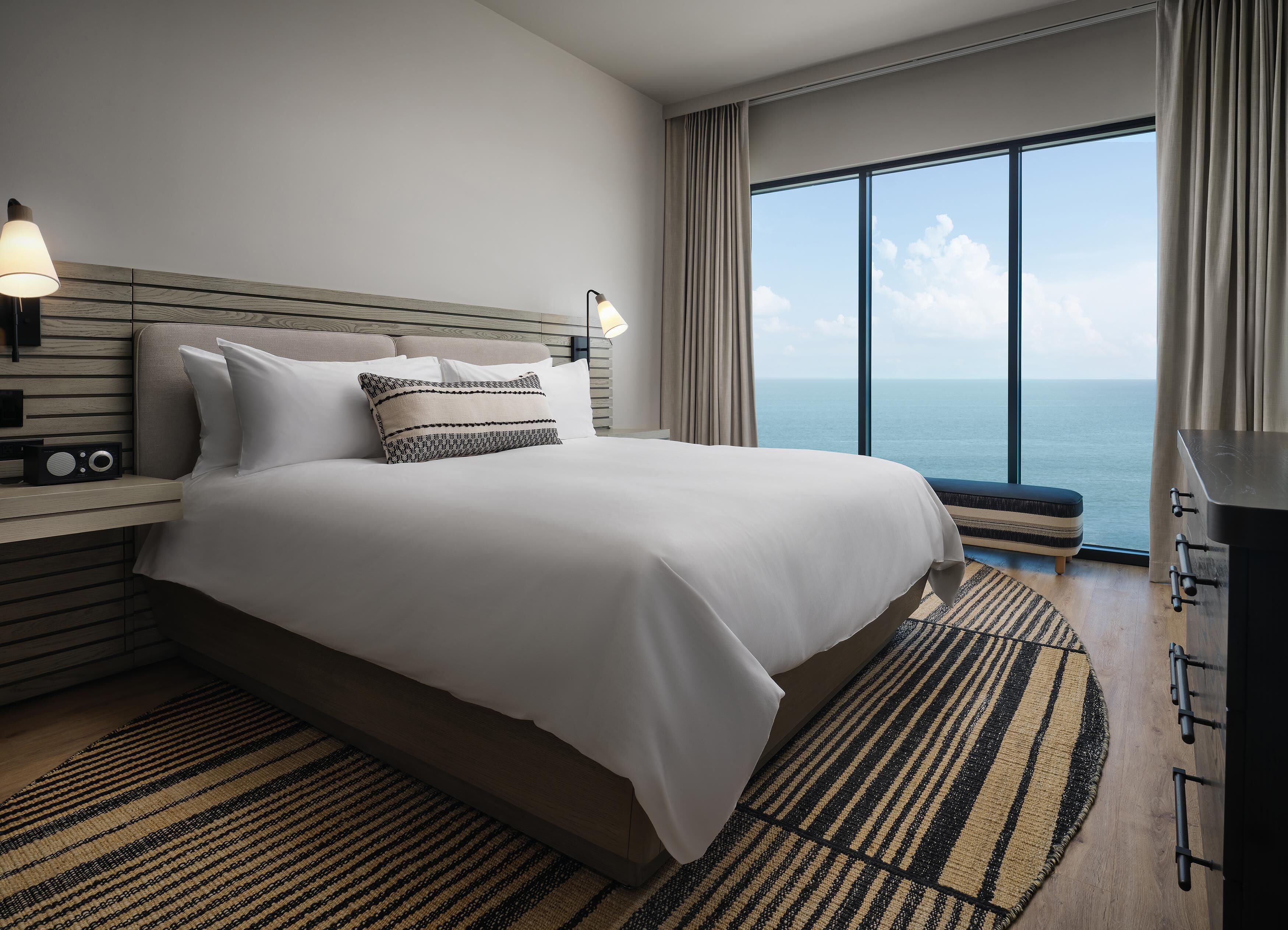 Un hotel moderno con una cama grande, mesitas de noche, una radio, vista al mar y un ventanal amplio.