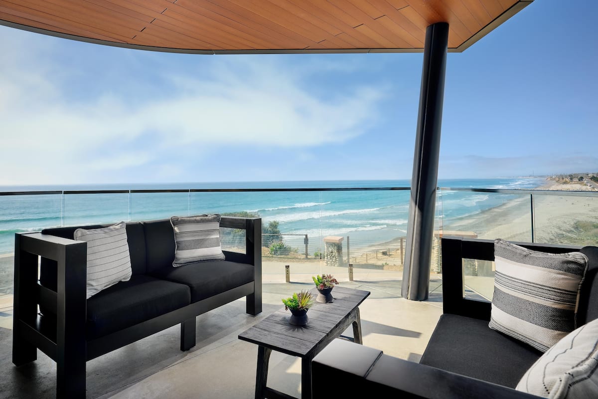 A balcony with a view of the ocean, a sofa, a small table with a plant, and a wooden ceiling.