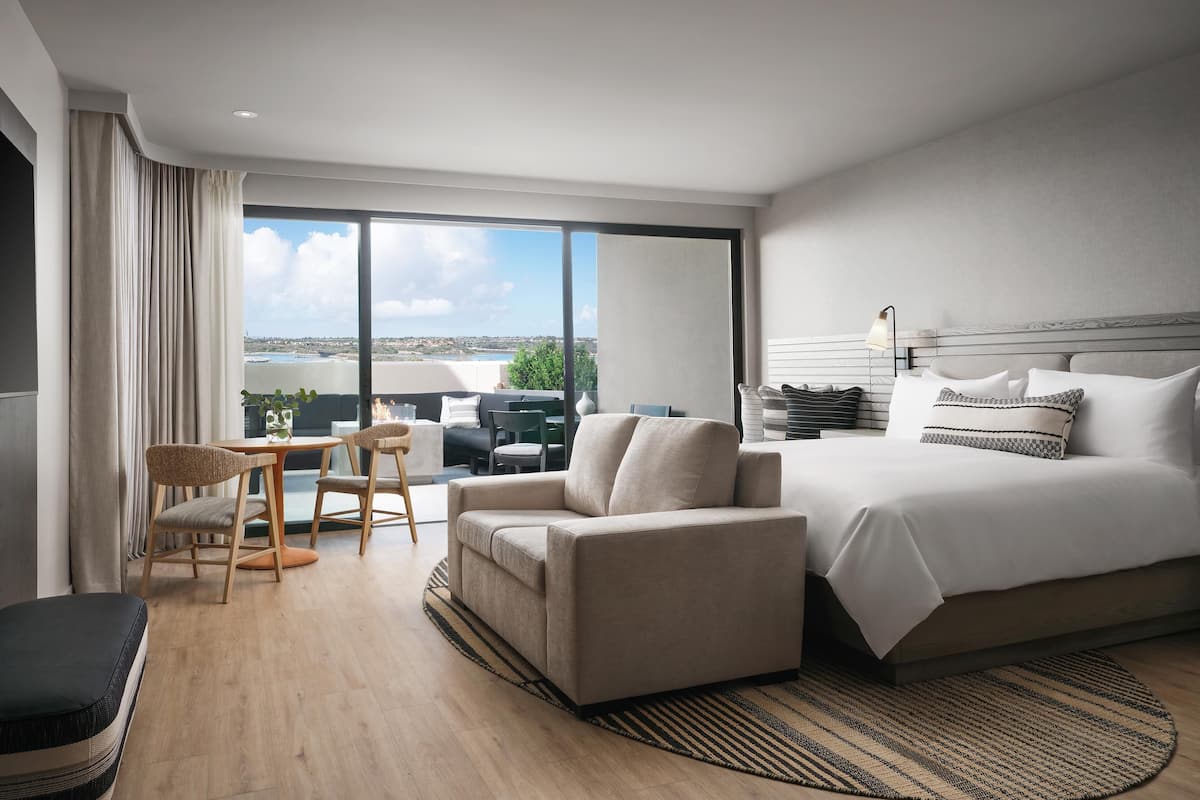 A modern hotel room with a large bed, a sofa, a dining area with chairs, and a view of the cityscape through large windows.