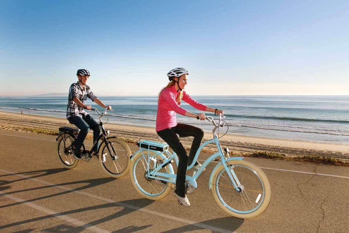 Two people riding bicycles on a paved path by the beach.