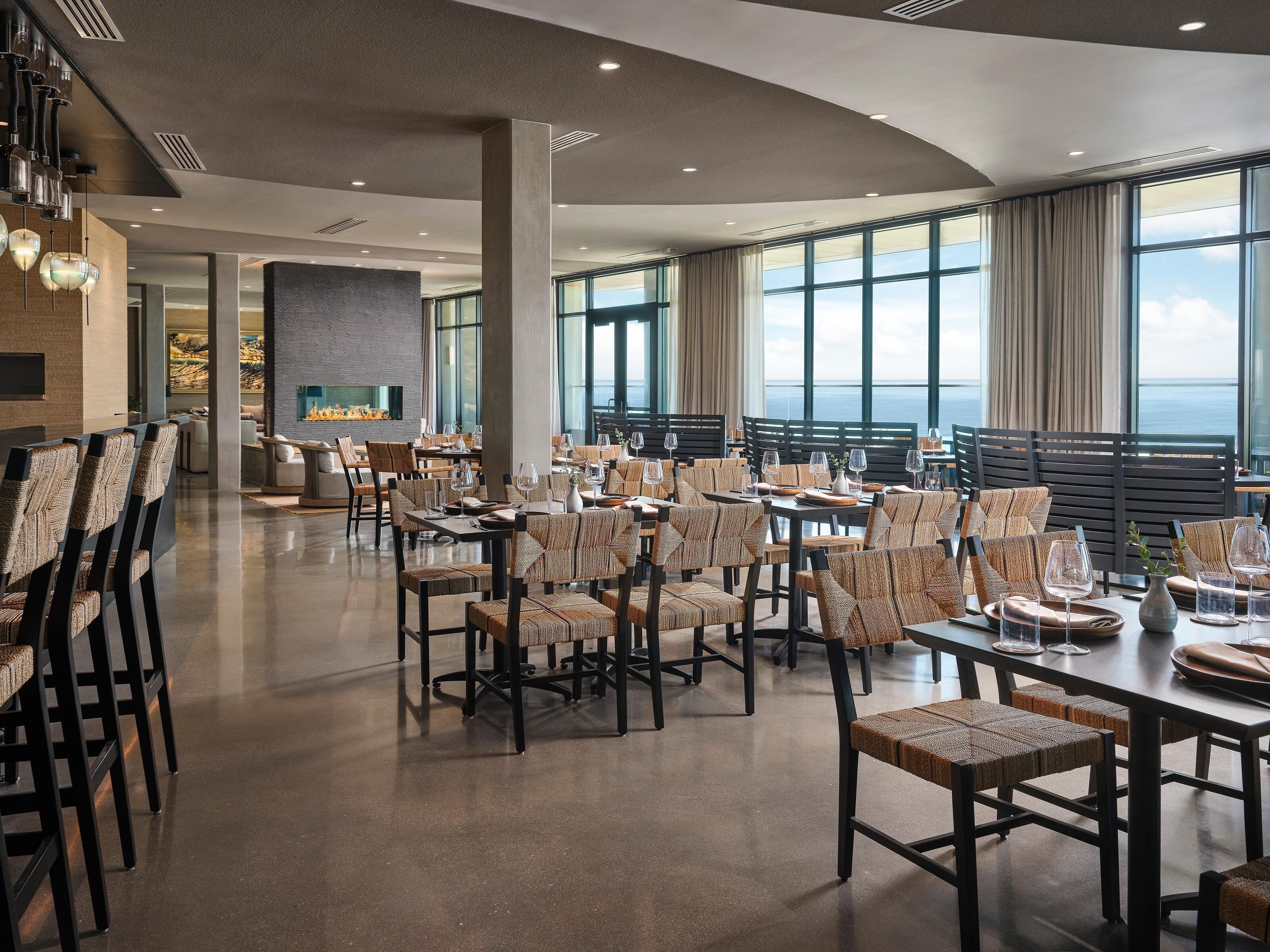 A spacious dining area with high tables and chairs, large windows, and a fireplace.