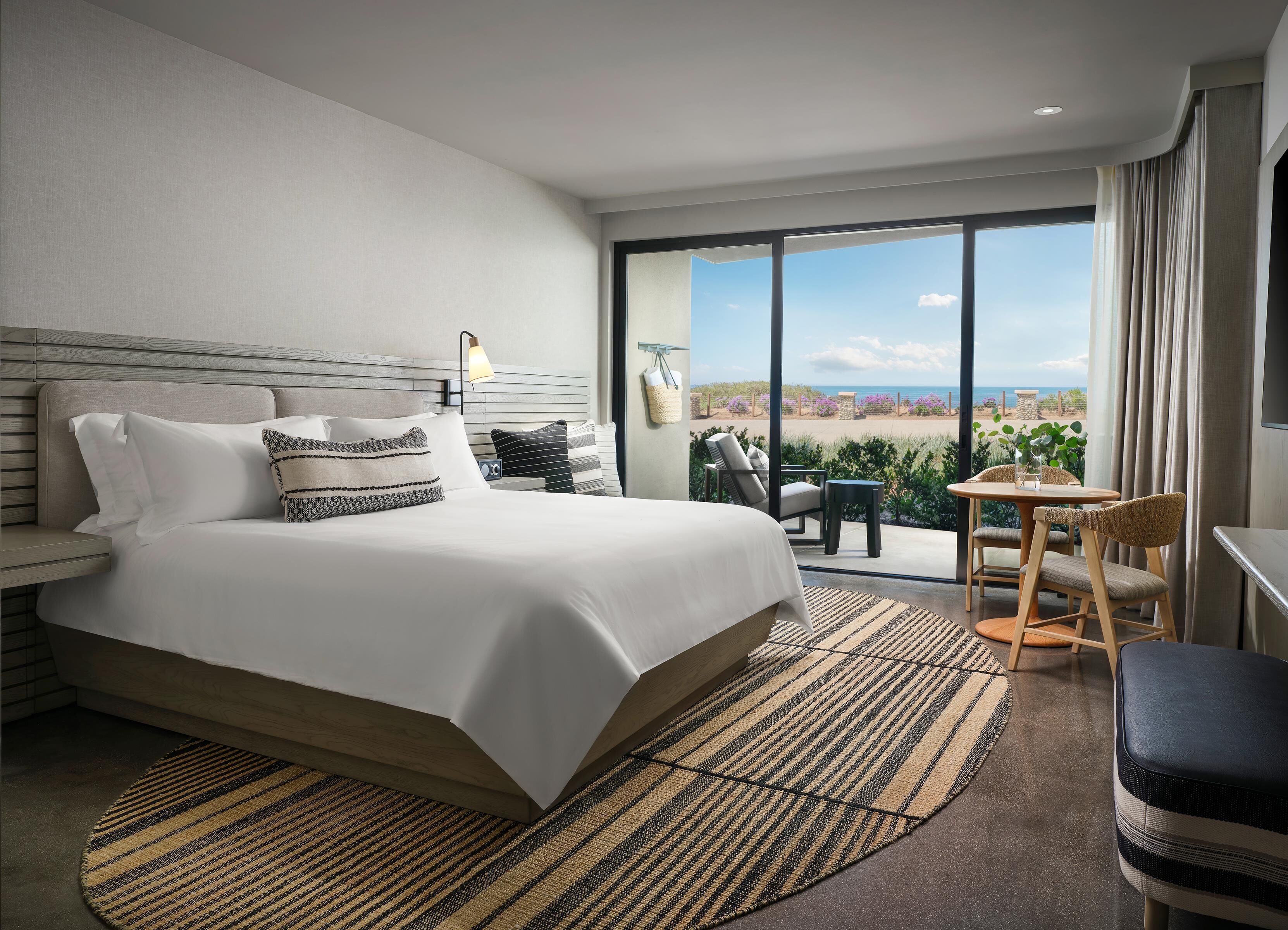 A modern hotel room with a large bed, a small table with chairs, and a balcony with a view.