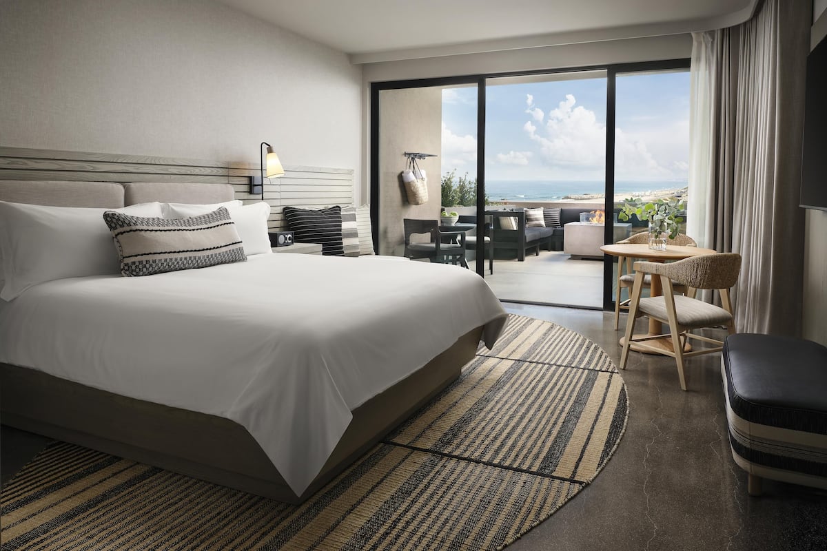 A modern hotel room with a large bed, a dining area with a table and chairs, and a balcony with a view of the ocean.