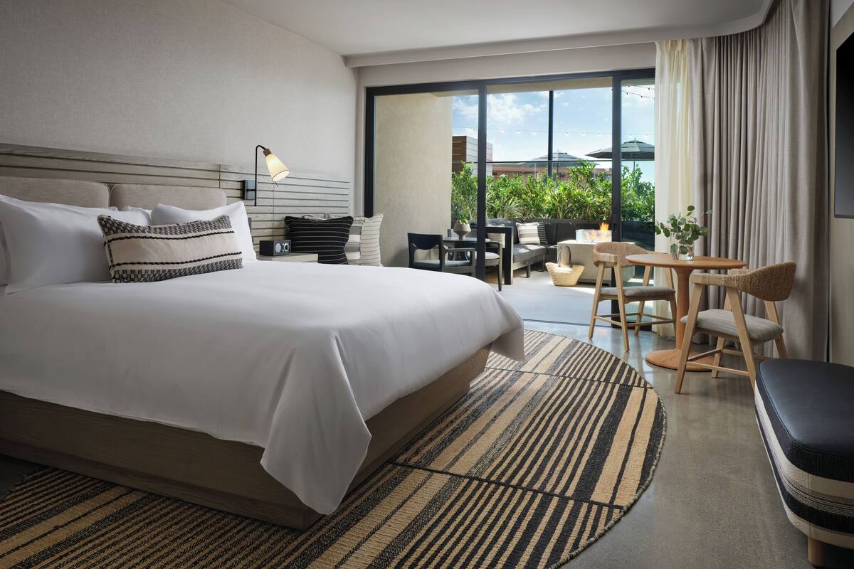 A modern hotel room with a large bed, a dining area, and a view of greenery outside.