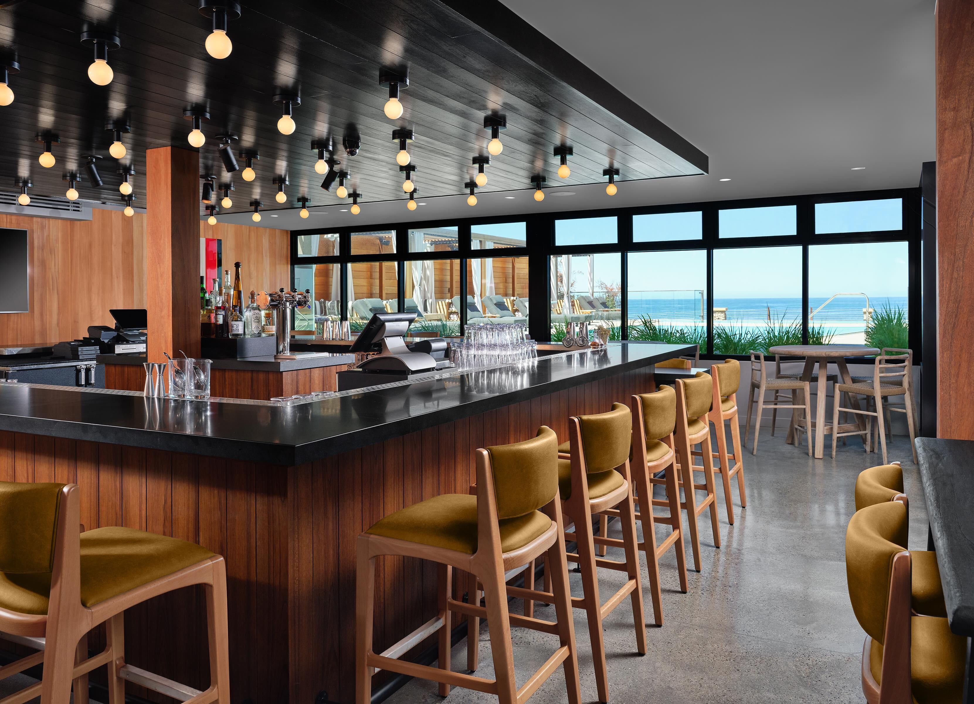 A modern bar area with a wooden bar counter, bar stools, and a view of the ocean through large windows.