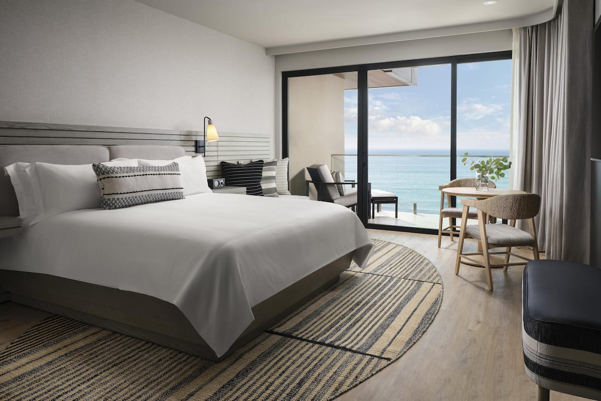 A modern hotel room with a large bed, a sitting area with a view, and a balcony overlooking the sea.