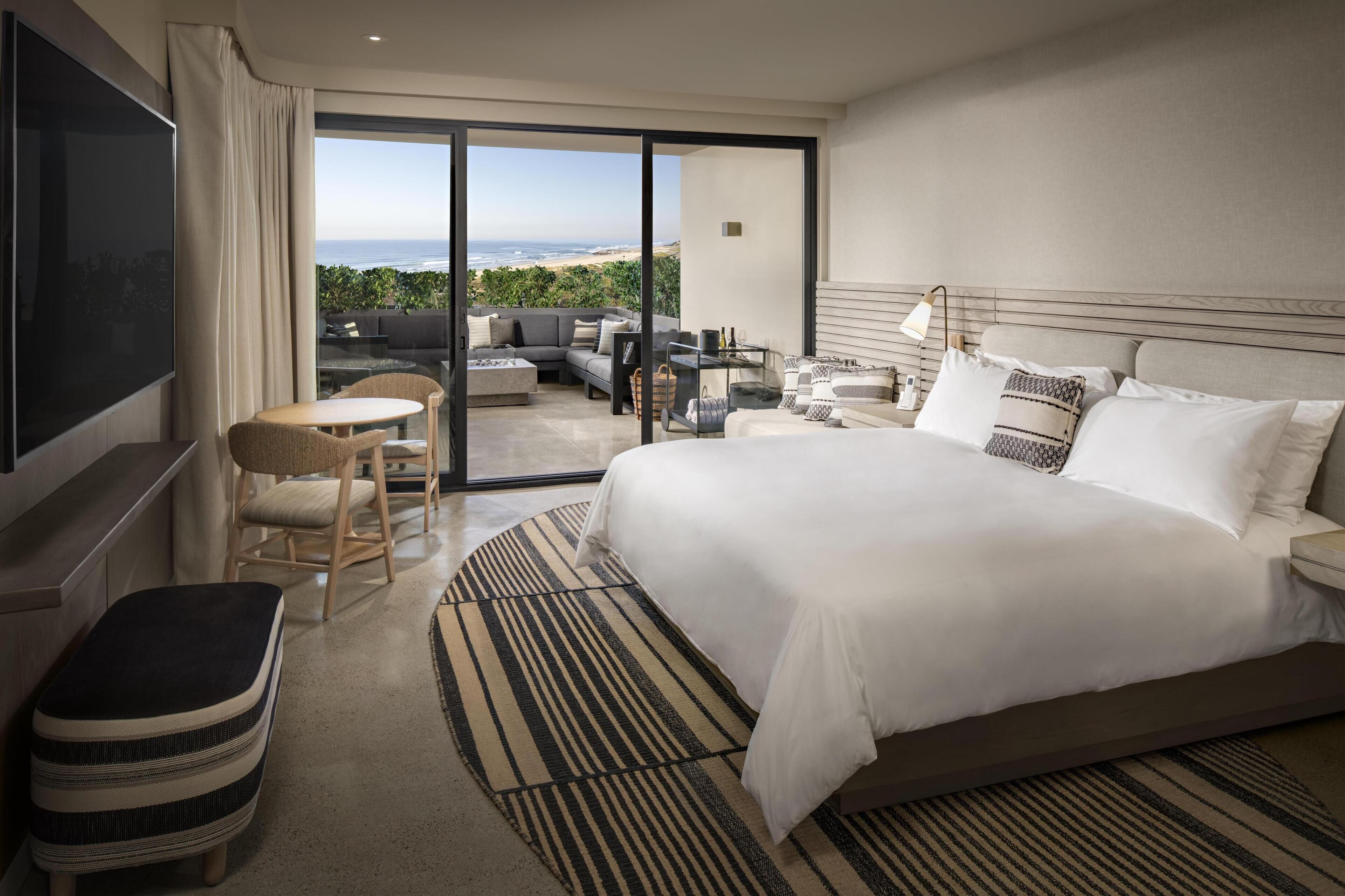 Habitación de hotel con una cama grande, televisor, zona de estar con mesa y sillas, y vistas al exterior.