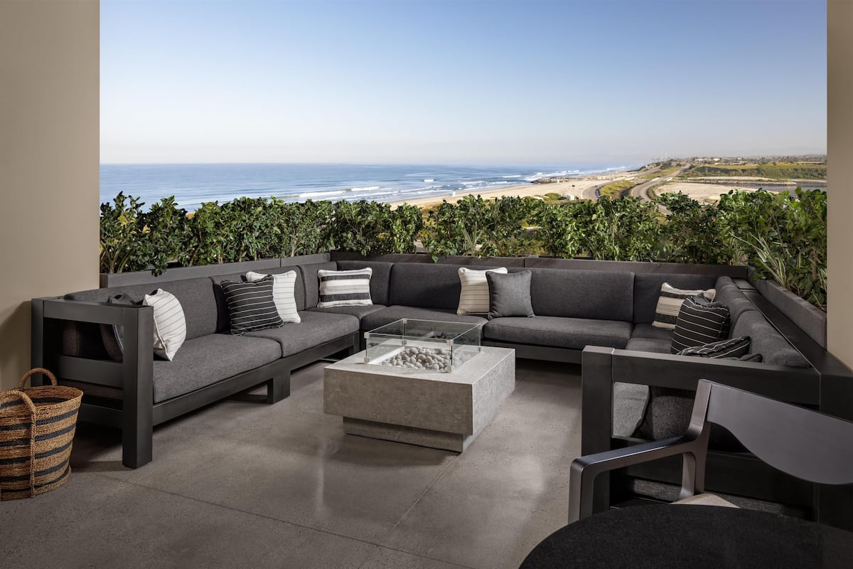 A modern outdoor seating area with a glass-top coffee table, grey sofas, and a view of the beach.