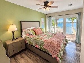 A bedroom with a bed, a nightstand, a lamp, a ceiling fan, and a view of the water through the balcony doors. - Luxury Waterfront Retreat with Private Dock, Golf Cart & Beach Access (Indian Rocks Beach)