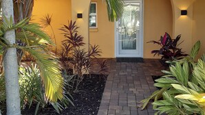 Un sendero conduce a una casa de color amarillo, rodeada de diversas plantas y un borde de ladrillo.