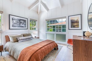 A bedroom with a bed, a desk, and a window with a view of a house.