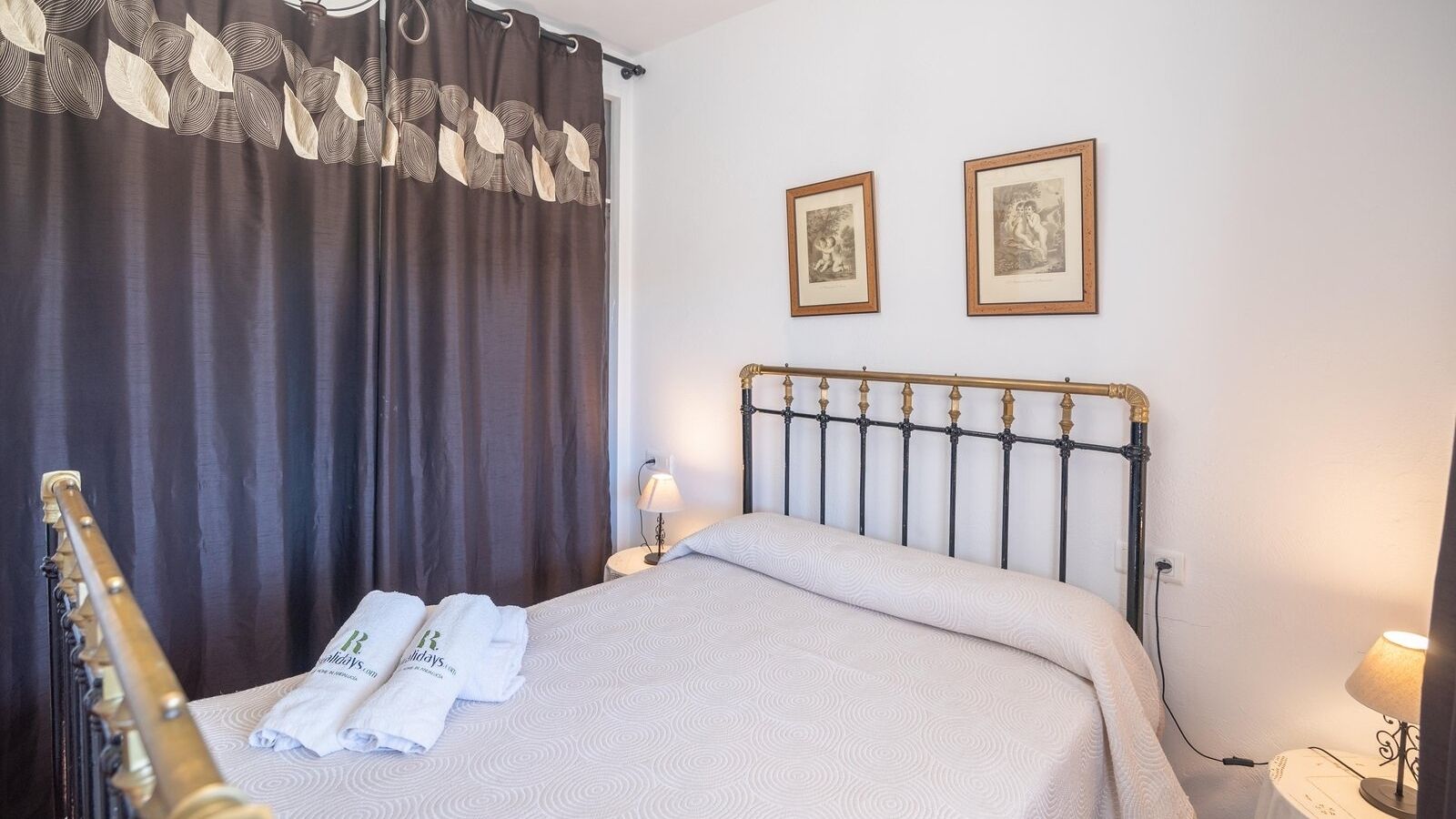 Una habitació d'hotel amb un llit, un capçal de llit, dues fotos amb marc a la paret, una làmpada i una guitarra recolzada contra la paret.