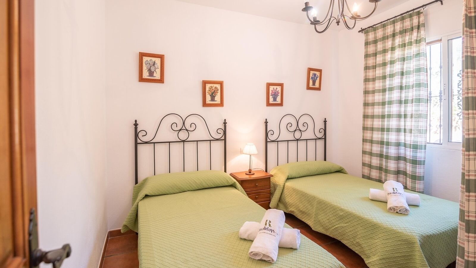 A room with two beds, each with green bedding and a rolled towel on the bed. There are framed pictures on the wall, a nightstand with a lamp, and a window with curtains.