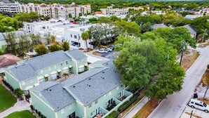 An aerial view of a residential area with a two-story light blue house with a grey roof, surrounded by greenery and a street.