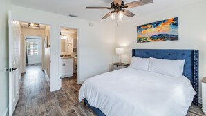 A bedroom with a large bed, a ceiling fan, a nightstand, a painting, and a hallway leading to a bathroom.