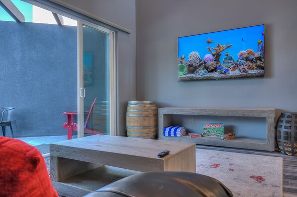 Een woonkamer met een televisie waarop een onderwaterlandschap te zien is, een houten salontafel, een rode stoel, een houten plank met een Monopoly-doos, en een glazen deur die naar buiten leidt.