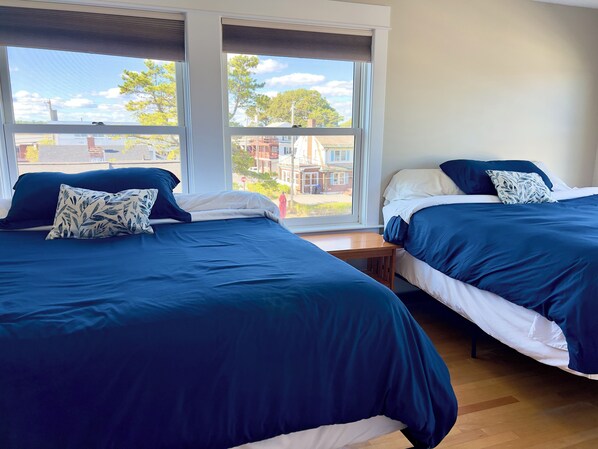 A room with two beds, each with a dark blue bedspread and matching pillows, a wooden nightstand, and large windows offering a view of buildings and trees.