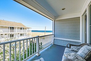 A balcony with a view of the beach, a white building in the background, and a wicker sofa with floral cushions.