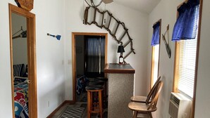 A spacious room with a wooden floor, a ceiling fan, a bar area with stools, and a bench.