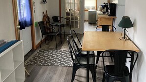 A cozy dining area with a wooden table and black chairs, a ceiling fan, and a view into a living room with a couch and a coffee machine.
