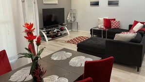 Een moderne woonkamer met een flatscreen televisie, een zwarte bank met rode en witte kussens, een eettafel met een rode plant, en een decoratief zeilschip op een bijzettafeltje.