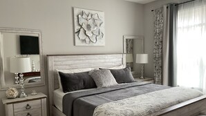Een slaapkamer met een houten hoofdbord, een groot bed met meerdere kussens, een nachtkastje met een lamp en een decoratieve spiegel.
