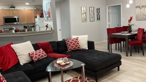 Een moderne woonkamer met een zwarte hoekbank, een glazen salontafel en een eethoek met rode stoelen.