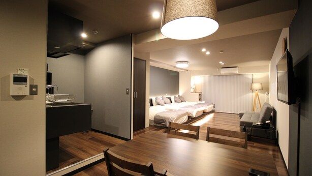 Una habitación de hotel moderna con una mesa de comedor de madera, una cama con cabecera blanca, un pequeño sofá y un televisor montado en la pared.