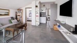 Una sala de estar moderna con una mesa de centro de vidrio, un sofá y una estantería. Hay una zona de cocina con un microondas y una lavadora.