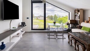 Una sala de estar moderna con un televisor de pantalla plana, un comedor con una mesa y sillas de vidrio, un ventanal con vistas a zonas verdes y una estantería con libros y un osito de peluche.