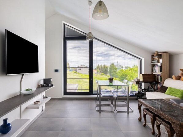 Una sala de estar moderna con un televisor de pantalla plana, un comedor con una mesa y sillas de vidrio, un ventanal con vistas a zonas verdes y una estantería con libros y un osito de peluche.