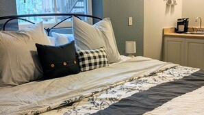 Een netjes opgemaakt bed met een bloemenprint sprei en een zwart-wit geruit kussen. Een raam met houten jaloezieën is zichtbaar, en een klok hangt aan de muur.