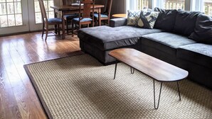 Een ruime woonkamer met een hoekbank, een eethoek met een tafel en stoelen, en grote ramen die voor natuurlijk licht zorgen.