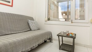 Ein Wohnzimmer mit grauem Sofa, einem gemusterten Kissen, einem kleinen Tisch mit einer Speiseplatte und einem Fenster mit Blick auf Gebäude.