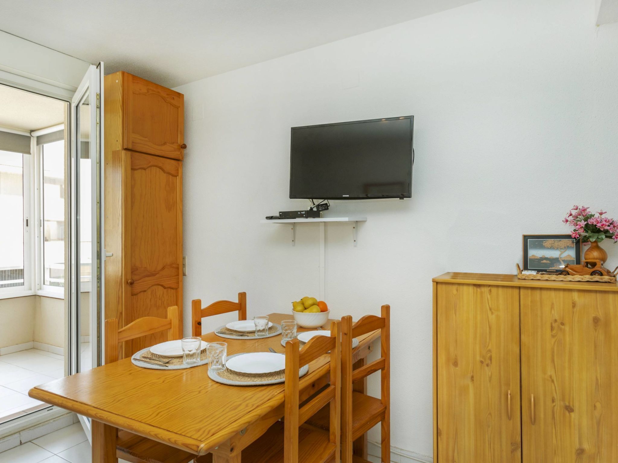 A dining area with a wooden table set for four, a television mounted on the wall, and a cabinet with a decorative item on top.