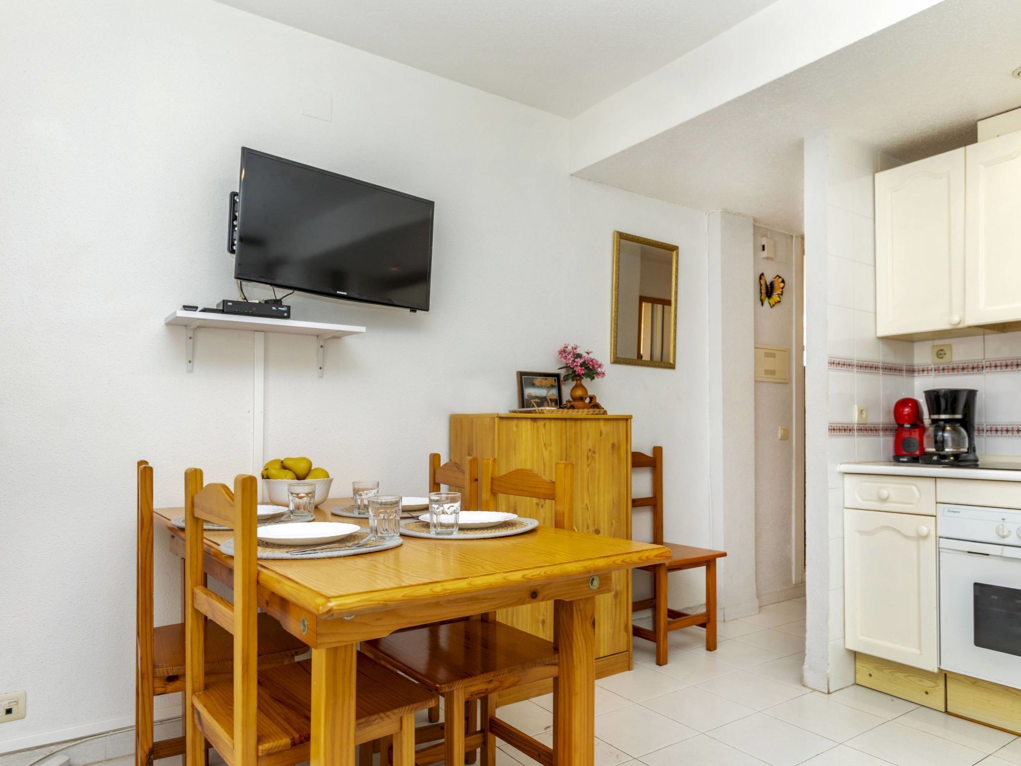 A dining area with a wooden table and chairs, a television mounted on the wall, and a kitchen area with white cabinets and a coffee maker.