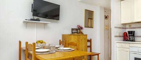 A dining area with a wooden table and chairs, a television mounted on the wall, and a kitchen area with white cabinets and a coffee maker.
