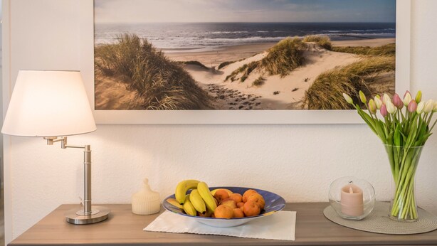 Un paysage de plage encadré, une table avec une lampe, un bol de fruits, une bougie et un vase de fleurs.