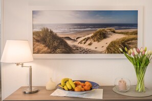 A framed beach landscape, a table with a lamp, a bowl of fruit, a candle, and a vase of flowers.