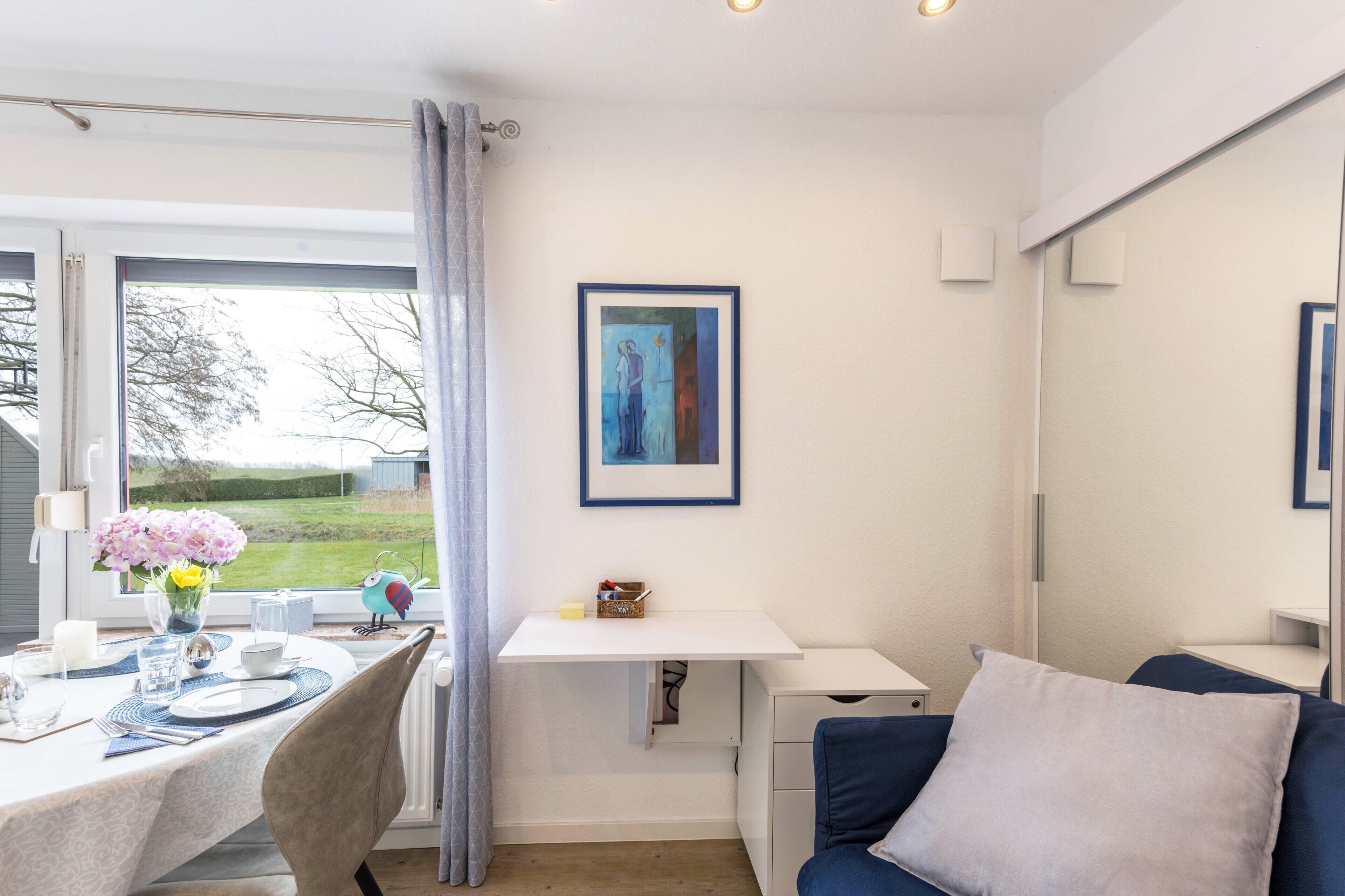 Ein Zimmer mit Esstisch für zwei Personen, einem kleinen weißen Schreibtisch, einem blauen Sofa und einem gerahmten Kunstwerk an der Wand.