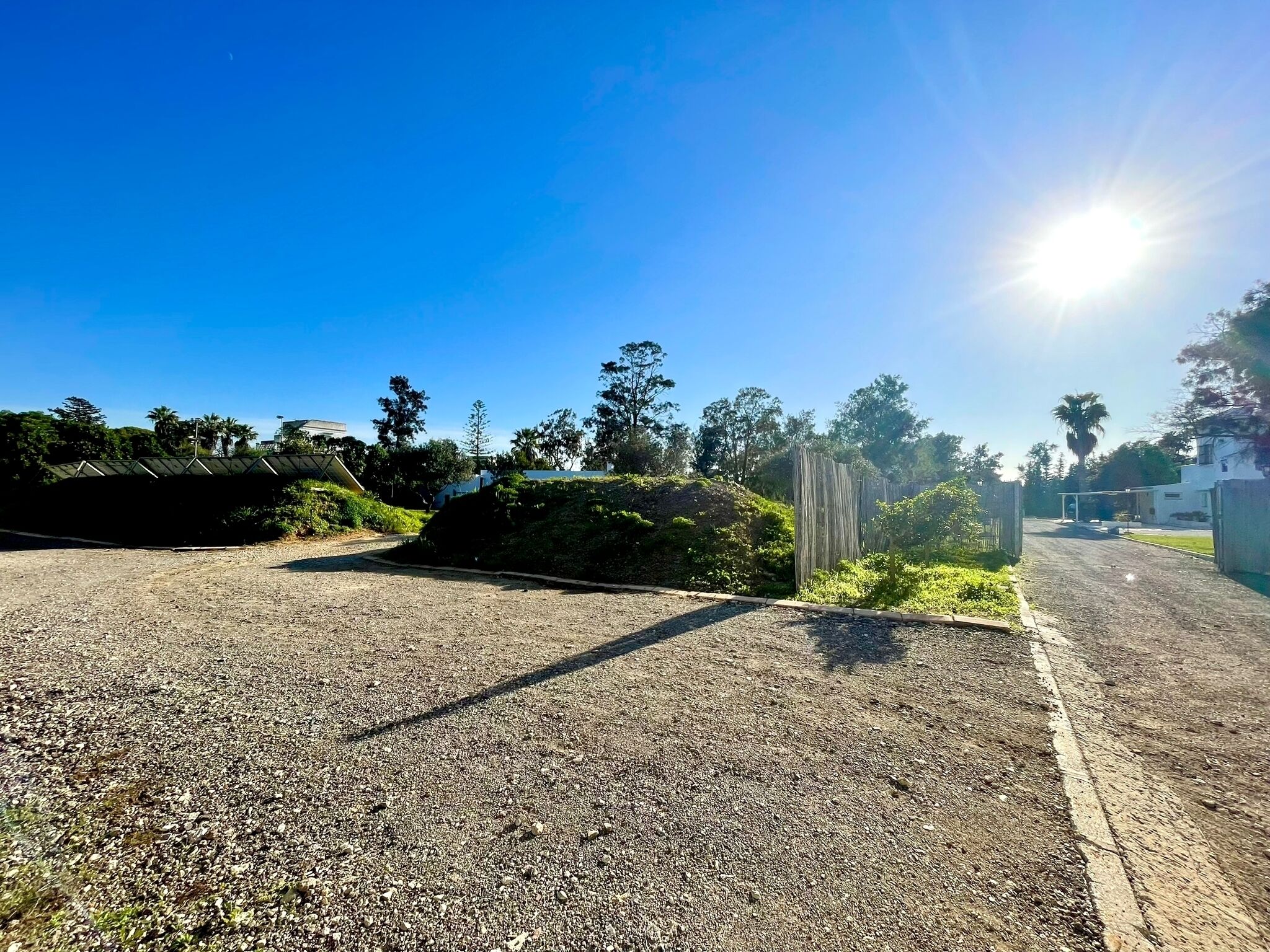 A gravel road with a metal bridge, surrounded by greenery and buildings.