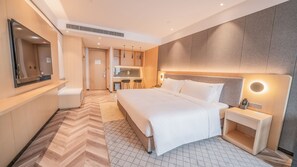 A modern hotel room with a large bed, a bedside table, a mirror, and a kitchenette area.