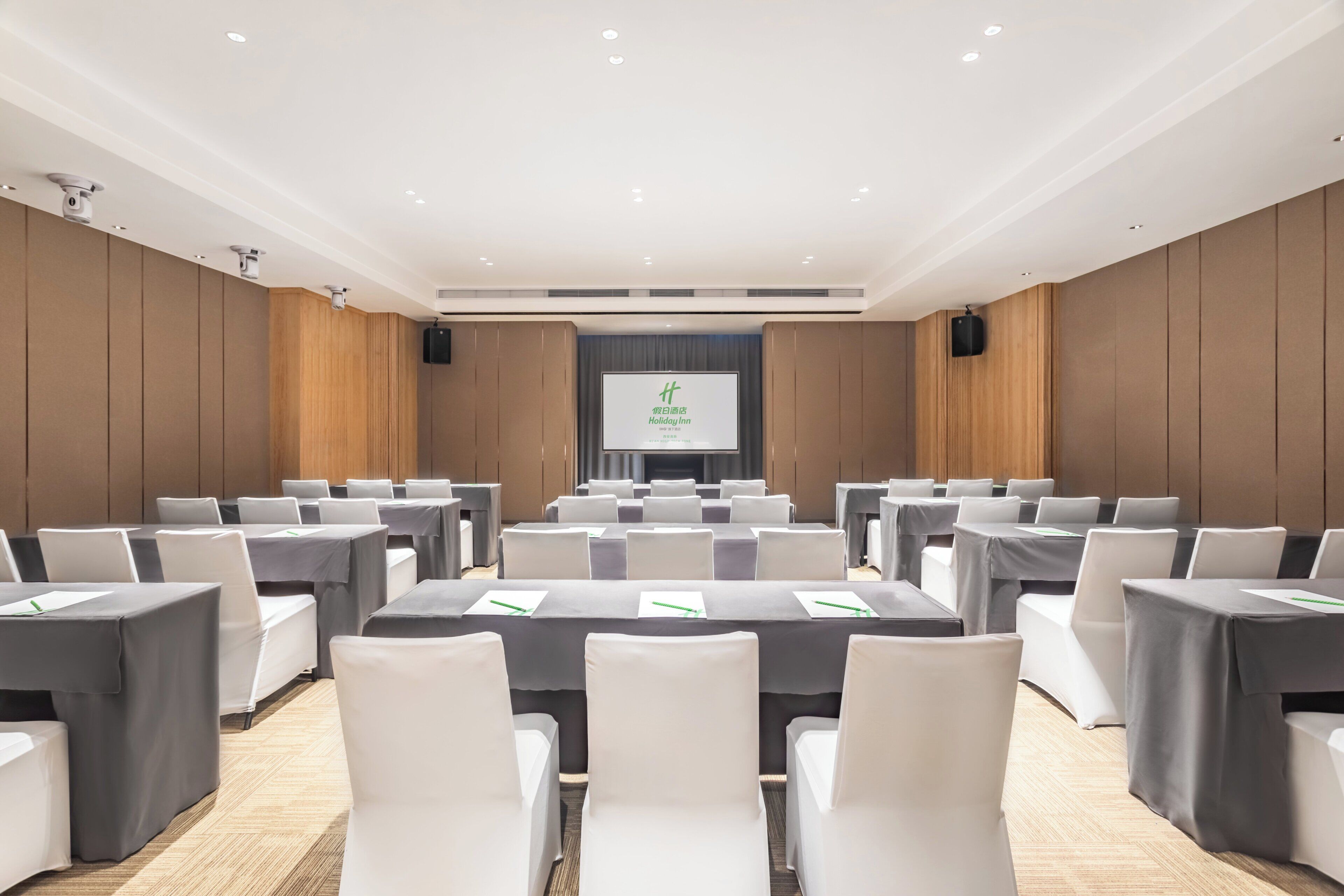 A conference room with rows of tables and chairs, a projector screen, and a wooden panel wall.