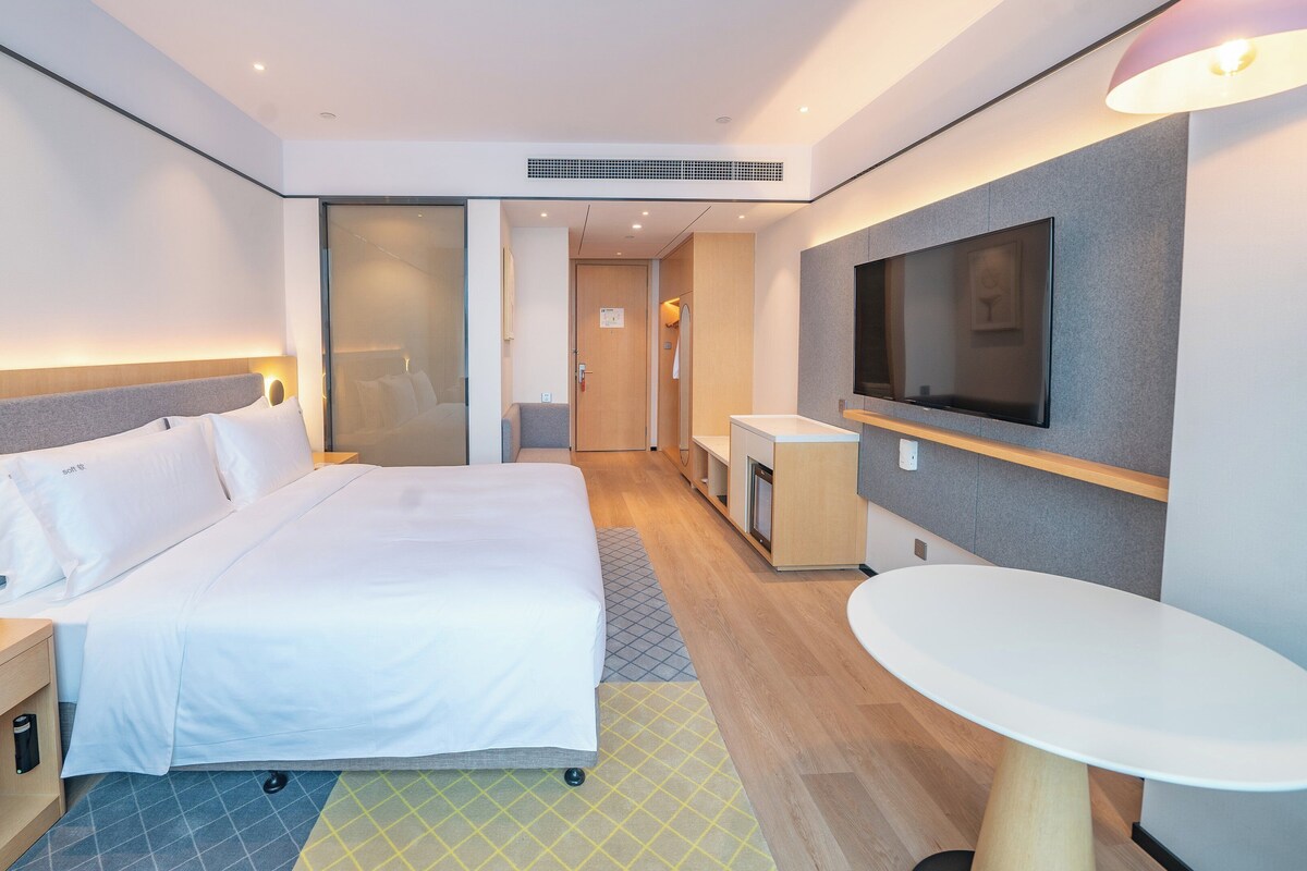A modern hotel room with a large bed, bedside table with a phone, a small desk with a chair, a TV mounted on the wall, and a small table.