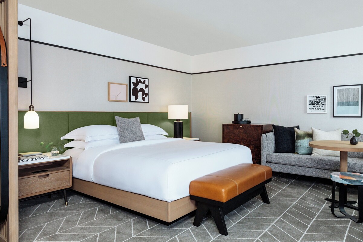 Een moderne hotelkamer met een groot bed, een slaapbank, een klein tafeltje en een bankje. Twee nachtkastjes met lampen, een groen hoofdeinde en kunst aan de muren.