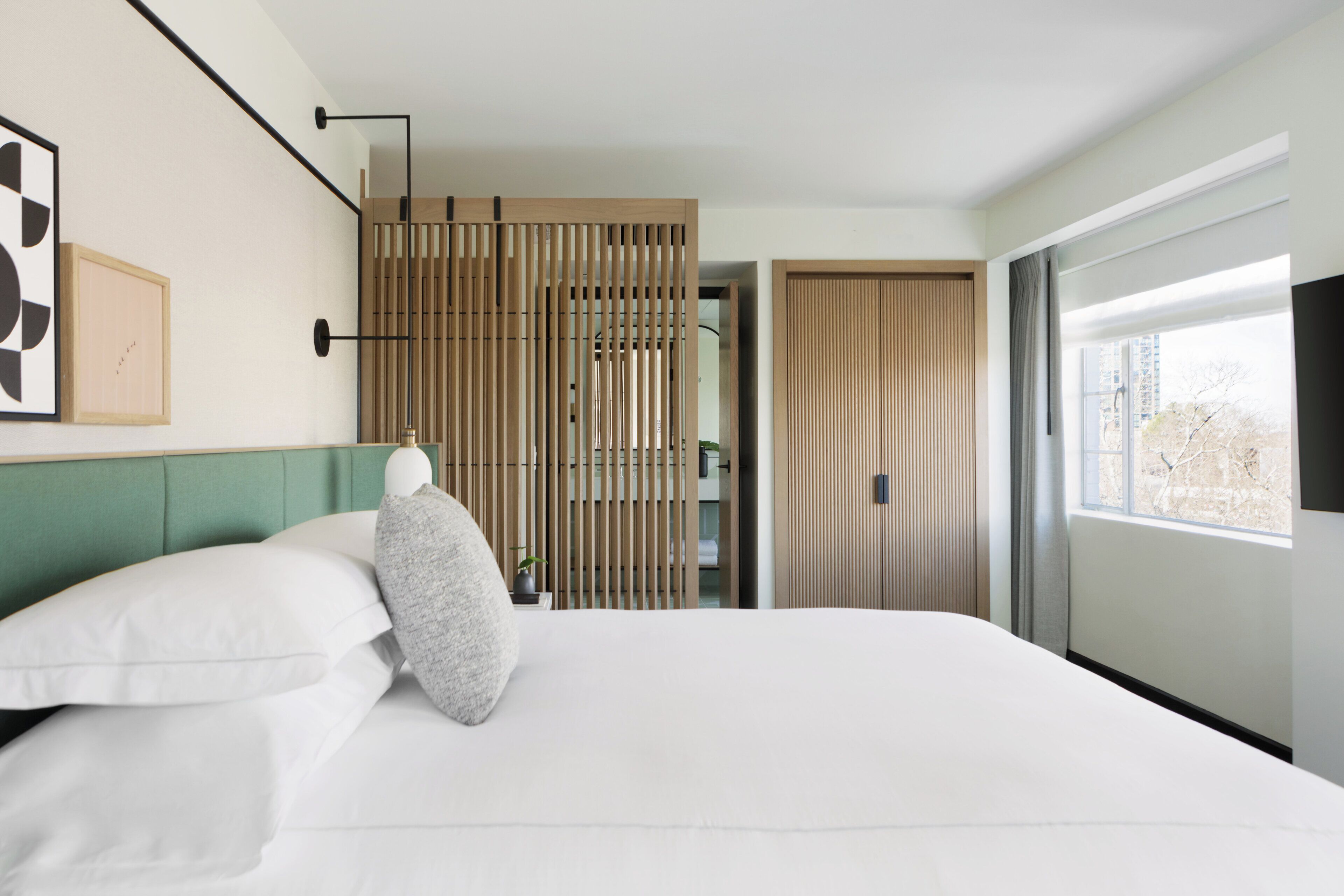 A modern hotel room with a bed, a view of the city through a window, and a sliding door leading to a bathroom.